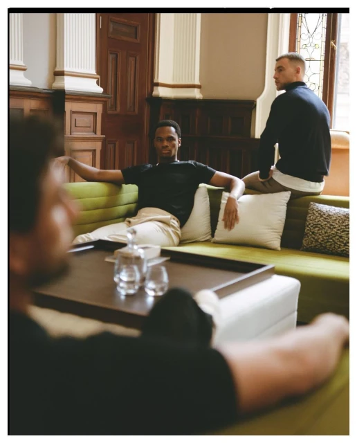 Three adult males relax indoors: one sits on the end of a green sofa, another leans back, and the third sits in the foreground.