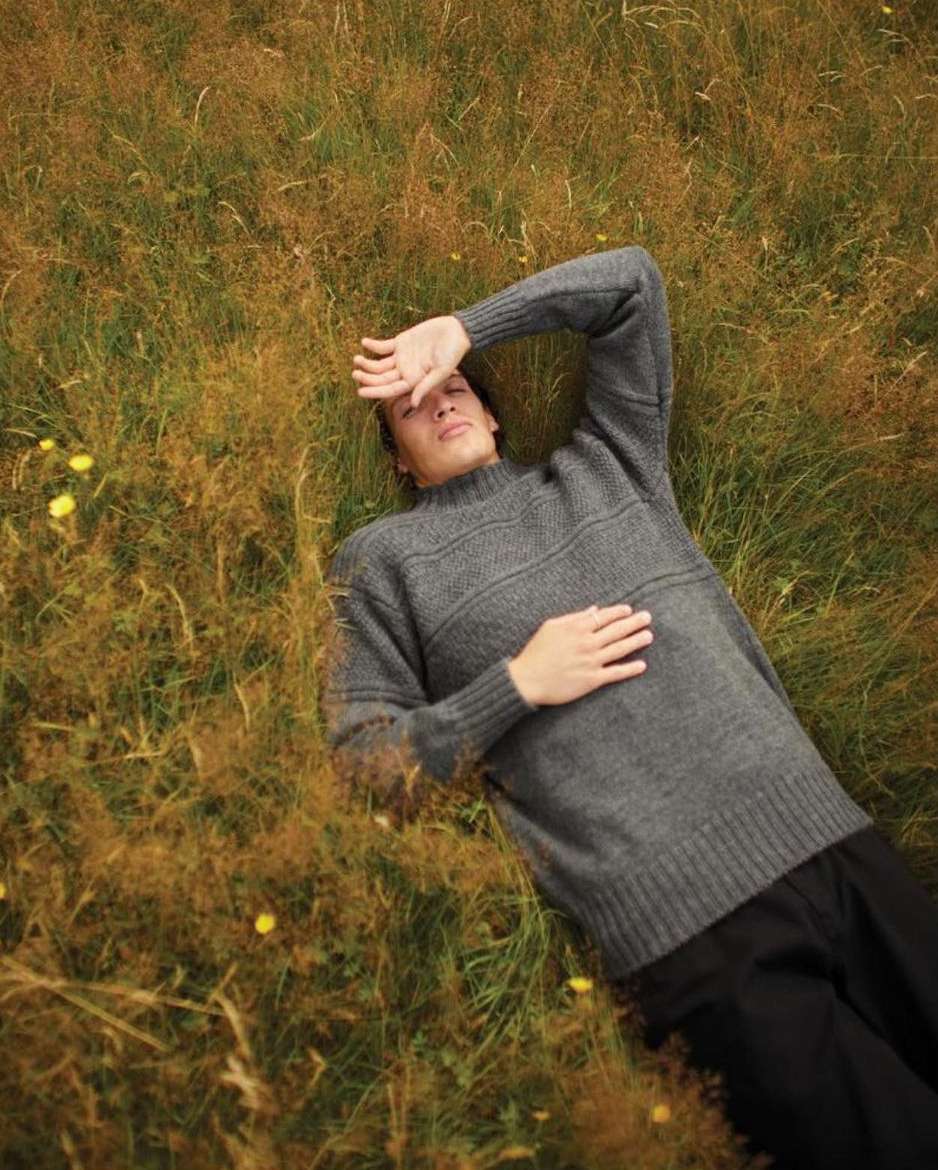 A fair-skinned adult male lies in tall grassy field, shielding his eyes in sunlight with a gray knit sweater, green and brown grass.