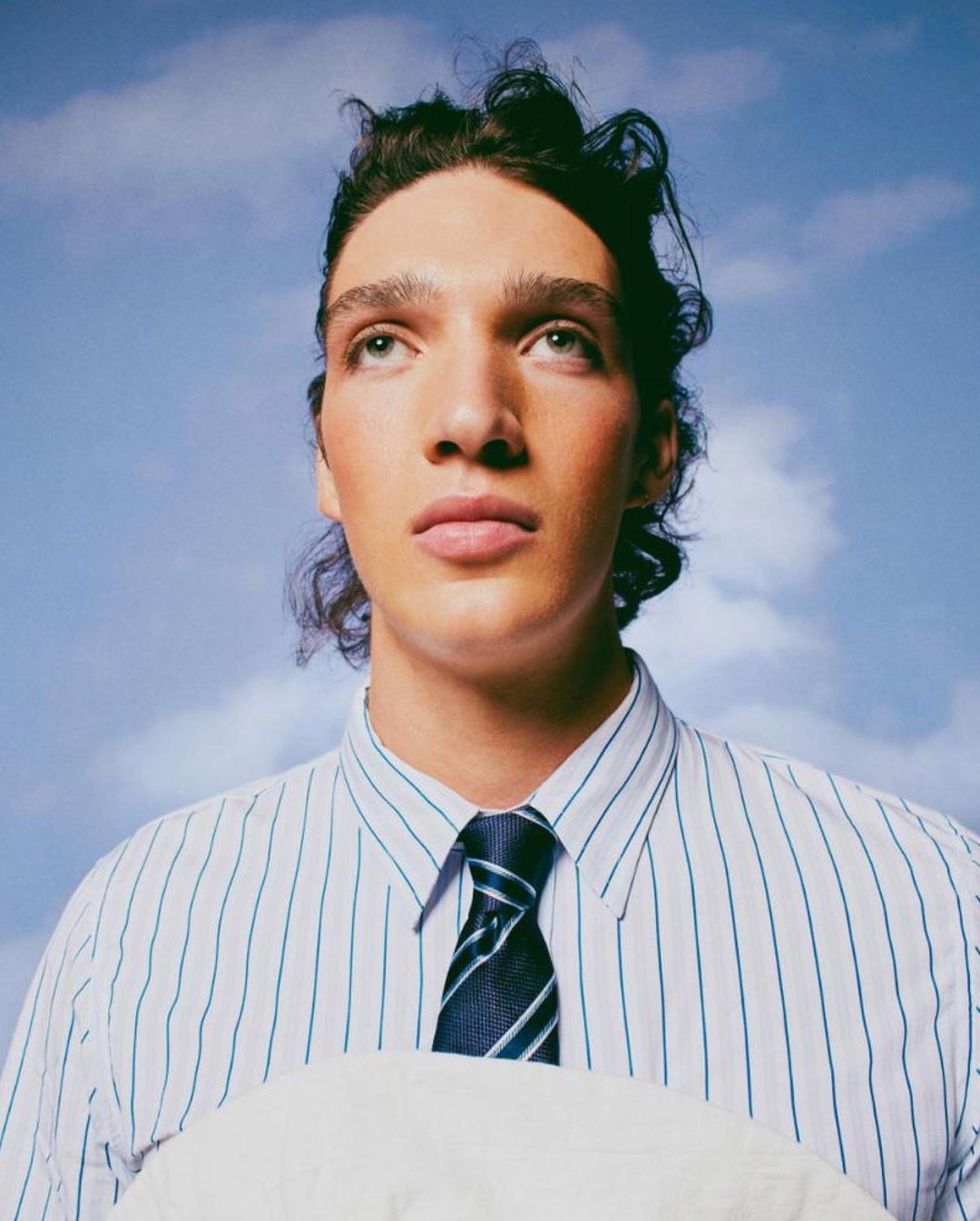 Portrait of a young adult male in a blue-and-white striped shirt and dark tie, holding a pillow, set against a blue sky with clouds.
