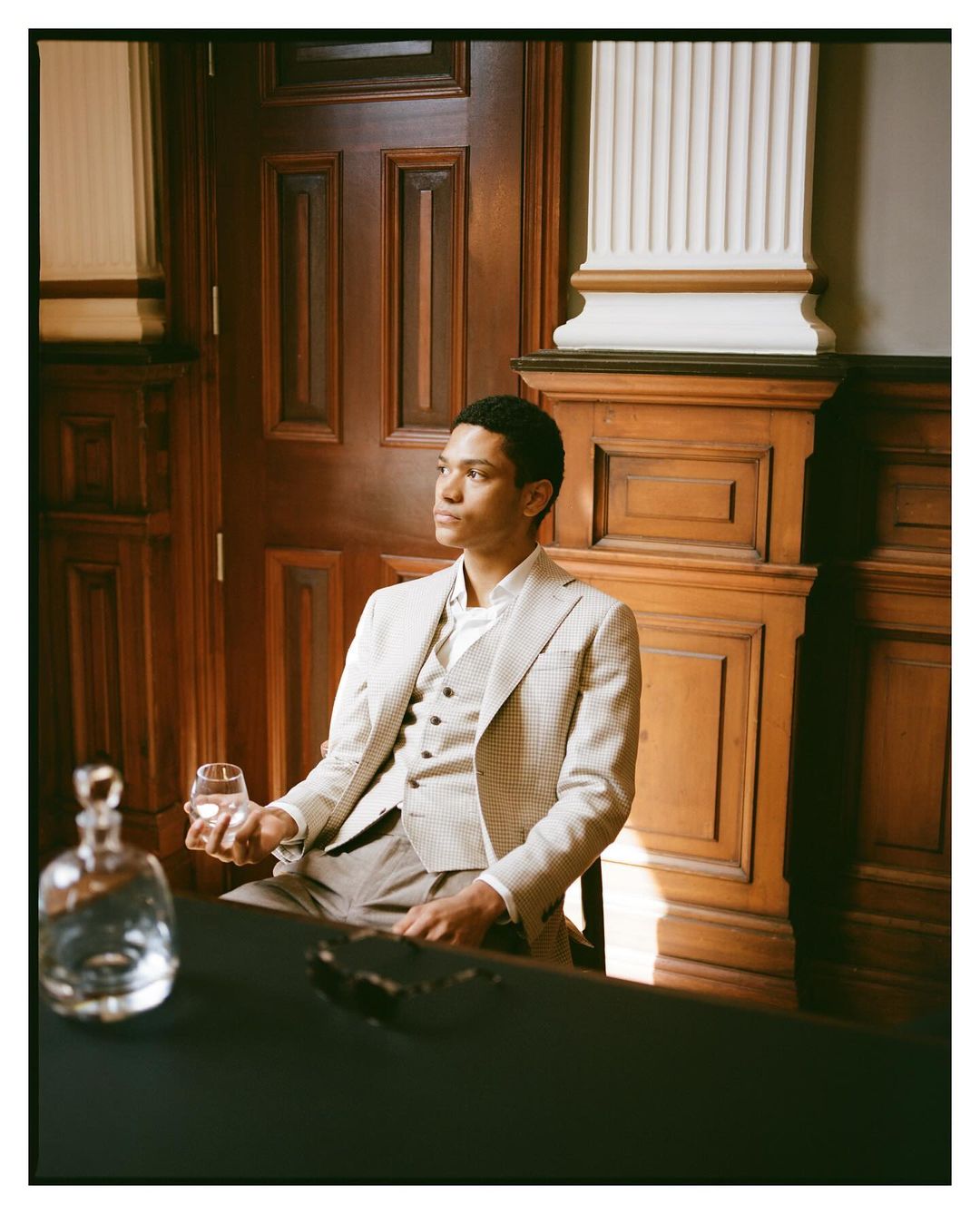 Well-dressed adult male sits indoors holding a glass. Dark table, wood paneling, natural light, with decanter and glasses nearby.