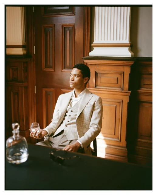 Well-dressed adult male sits indoors holding a glass. Dark table, wood paneling, natural light, with decanter and glasses nearby.
