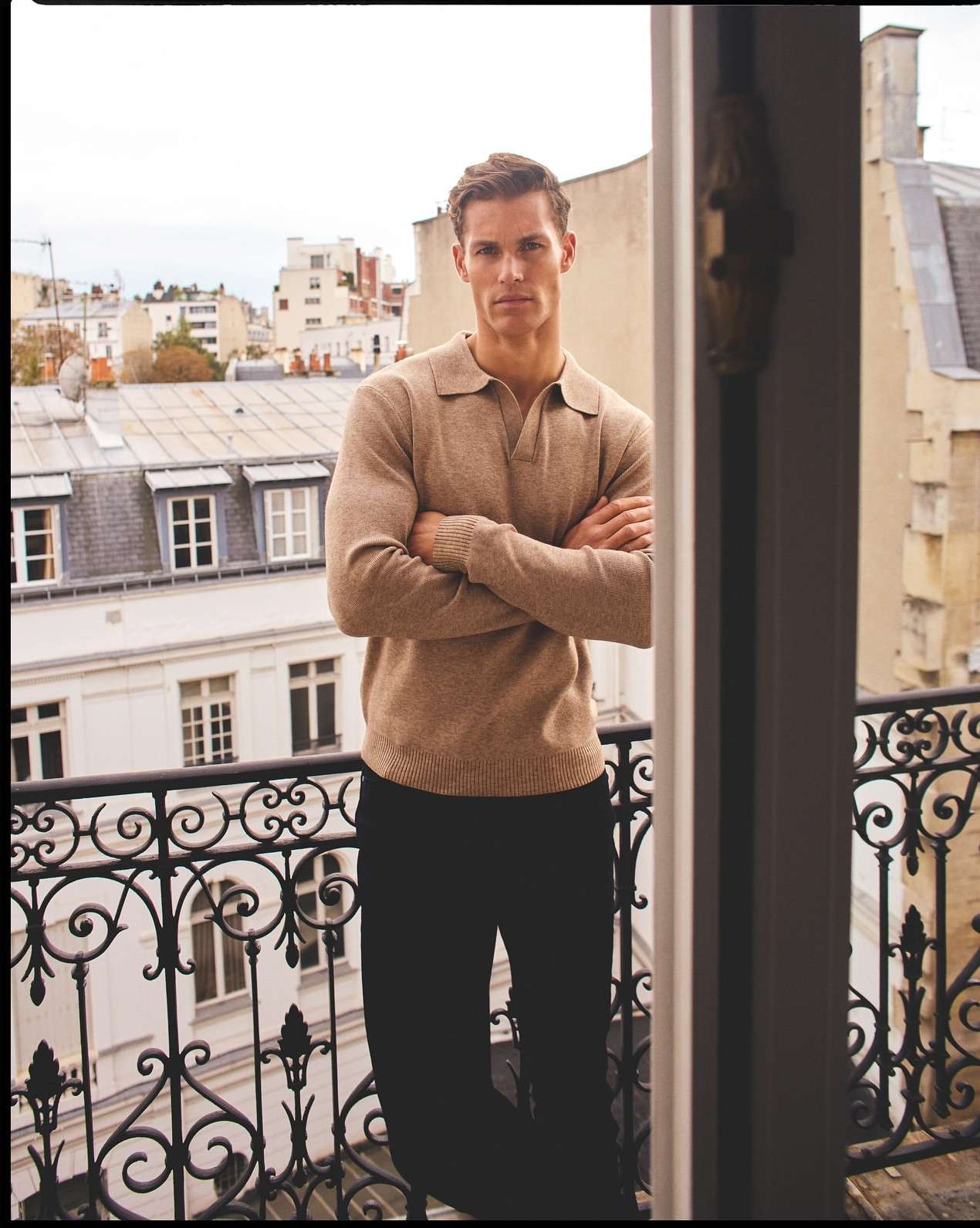 Handsome man stands on balcony in casual attire, gazing forward against a backdrop of Parisian rooftops and a cloudy sky, fashion shot.