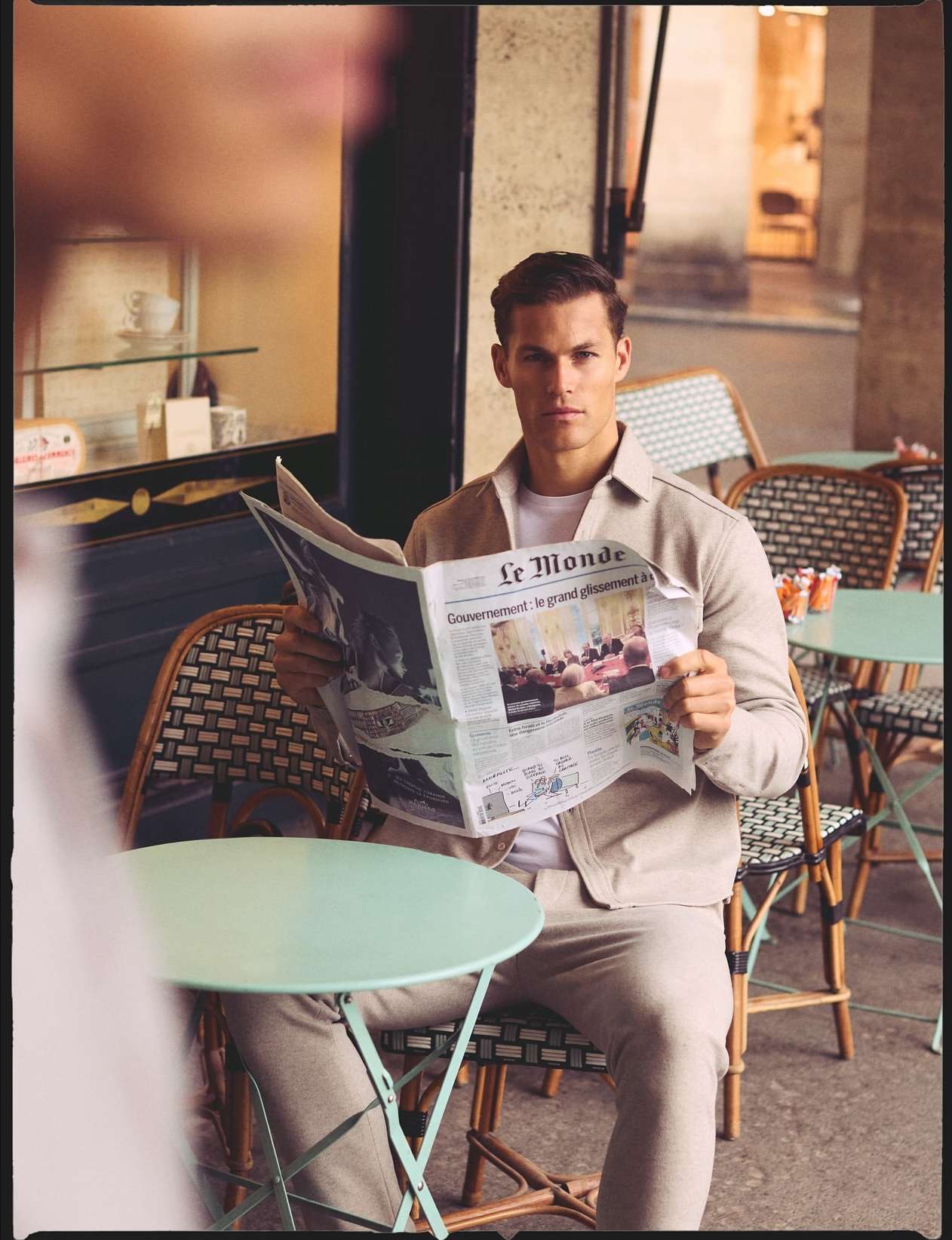 An adult male is sitting outdoors reading a newspaper in a cafe setting, wearing a neutral colored jacket and pants. The background shows an urban environment.