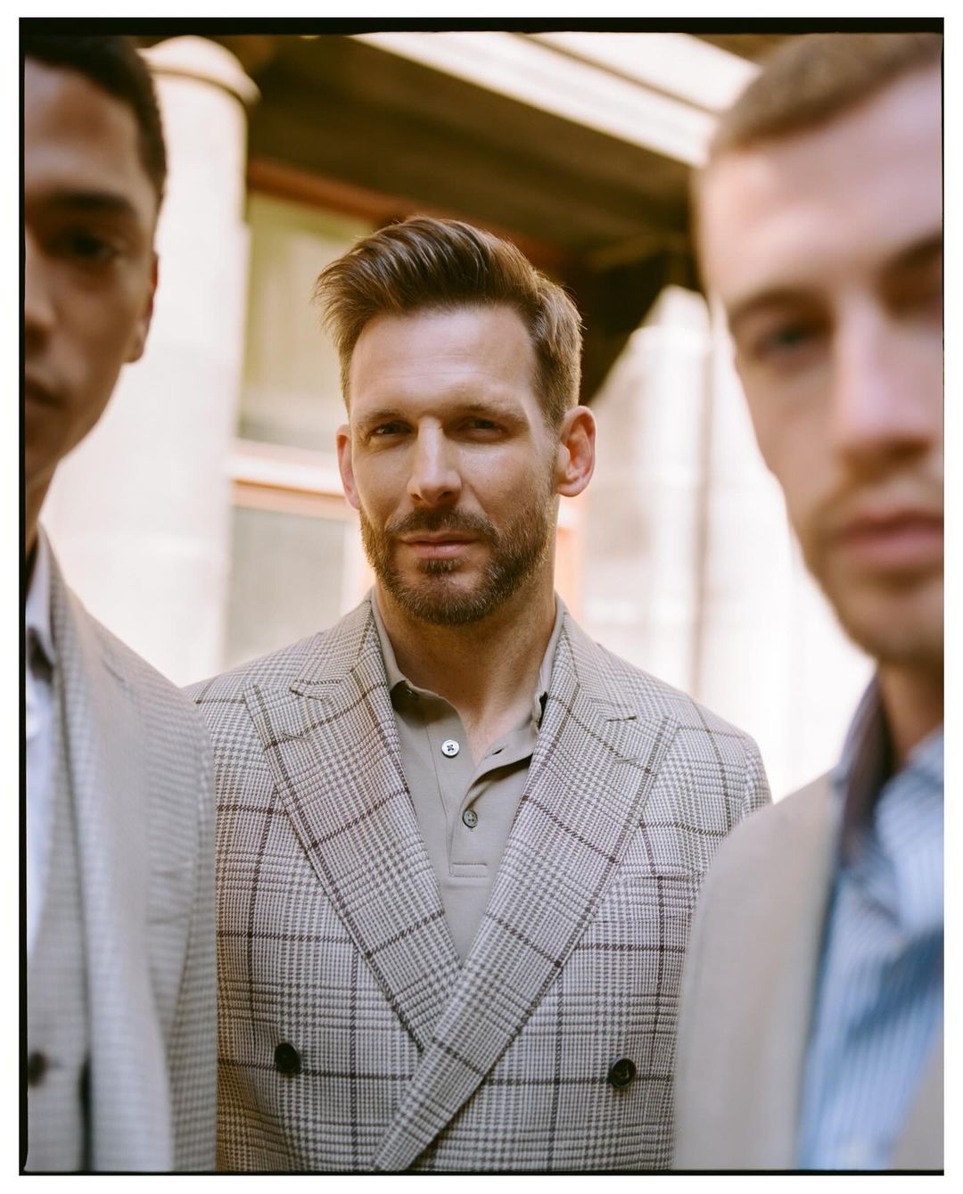 Three adult men pose outdoors wearing suits and polo shirts. The man in the middle has a beard and his hair is neatly styled.