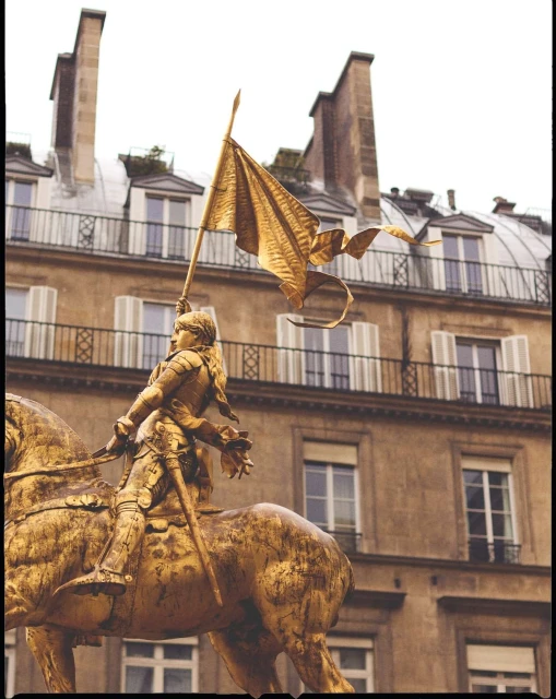 Golden statue of Joan of Arc on horseback holding a flag, set against an urban backdrop with classic European architecture.