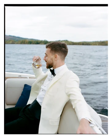 Man in formal attire on a boat, sipping a drink with lake and tree background under a cloudy sky, exuding a sophisticated and relaxed vibe.