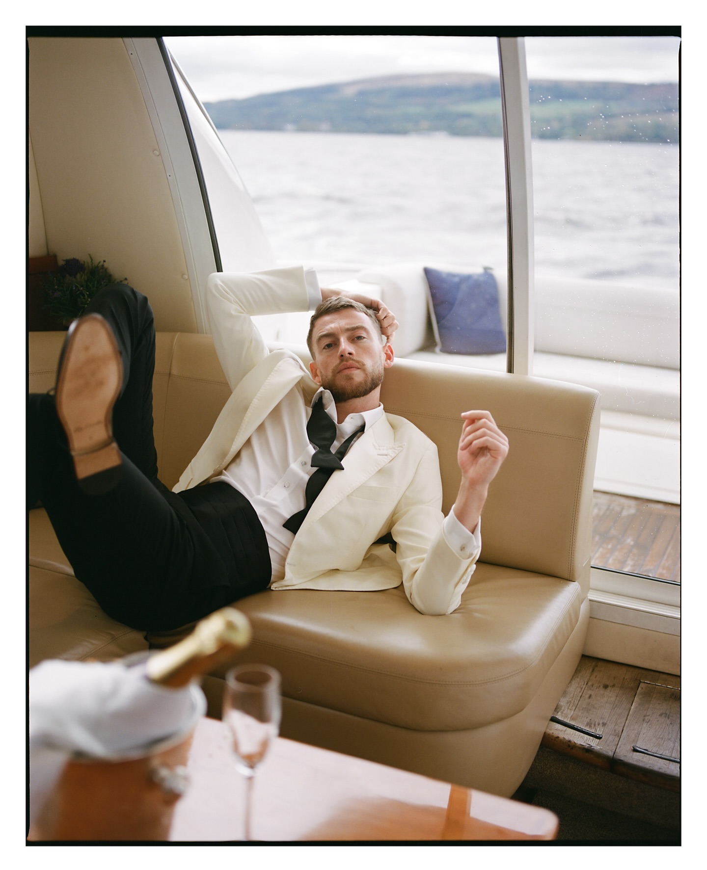A casually posed male model in a white tuxedo lounges on an indoor boat couch with champagne and water view, creating a lifestyle fashion editorial.