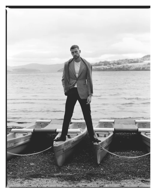 Black and white outdoor portrait of a man in a blazer standing on several canoes near a lake with mountains in the distance.