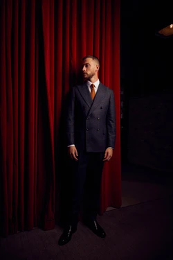 Man in double-breasted suit stands confidently against a red curtain in a dimly lit setting, creating a striking contrast.