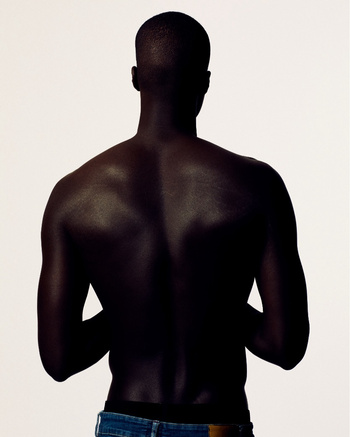 Back view of a dark-skinned adult male with a shaved head wearing blue jeans, in a studio setting with a bright white background.