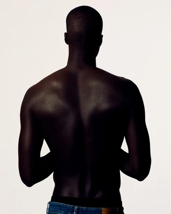 Back view of a dark-skinned adult male with a shaved head wearing blue jeans, in a studio setting with a bright white background.