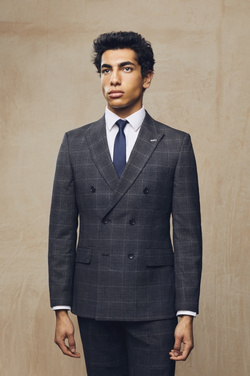 A man wearing a gray double-breasted suit, white shirt, and navy tie strikes a pose indoors with a neutral, textured backdrop.