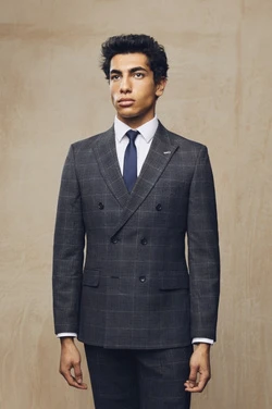 A man wearing a gray double-breasted suit, white shirt, and navy tie strikes a pose indoors with a neutral, textured backdrop.