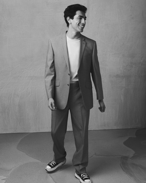 Man wearing a tailored suit jacket over a t-shirt and trousers with sneakers, smiling in a studio setting