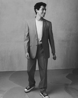 Man wearing a tailored suit jacket over a t-shirt and trousers with sneakers, smiling in a studio setting
