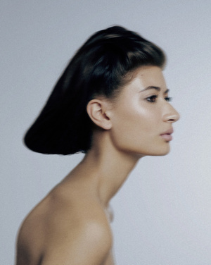 Profile view of a brunette adult female model with slicked-back hair against a pale blue gradient background in a beauty shot.