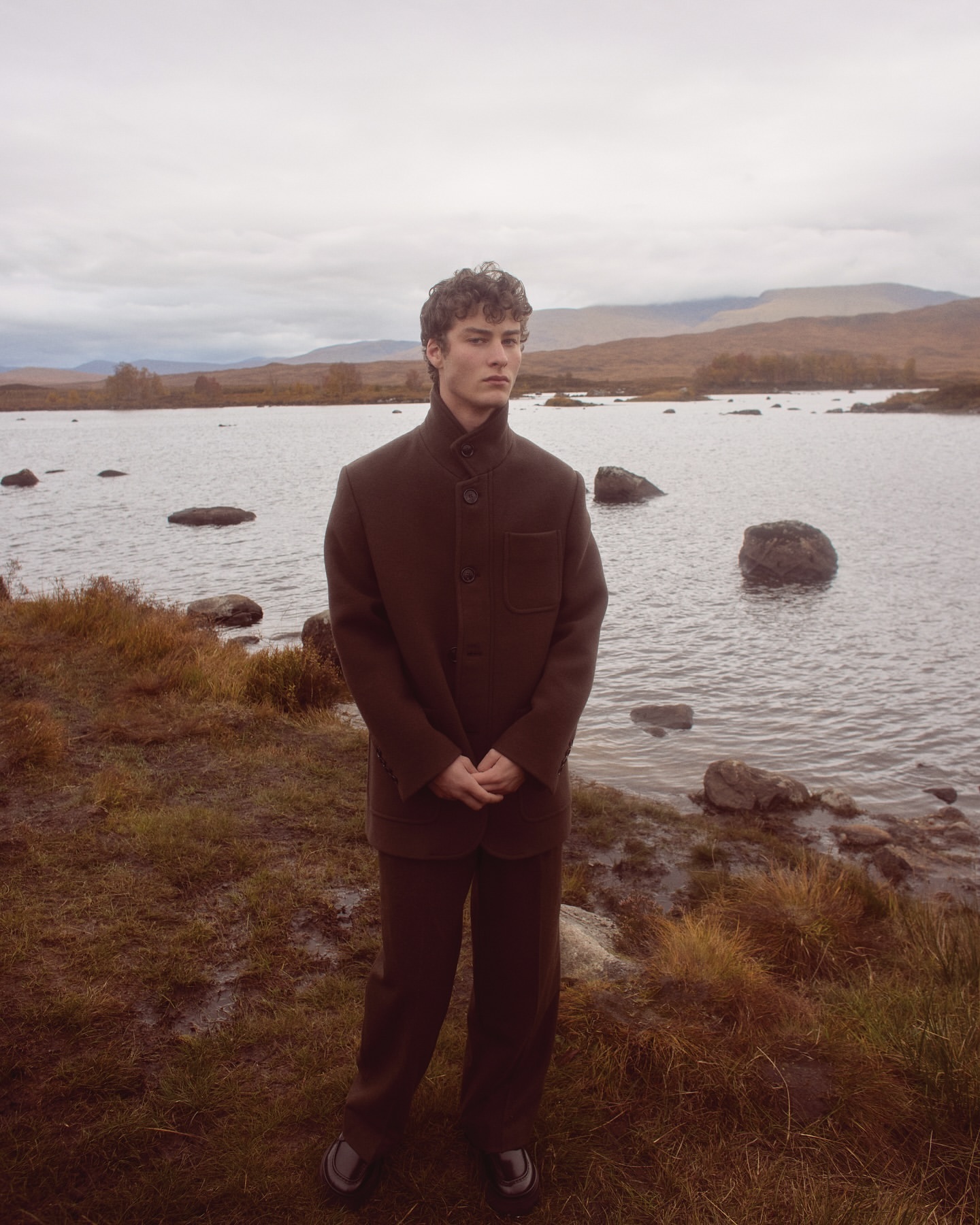 Young man in brown suit stands by a lake with mountains in the background under an overcast sky. Fashion shot, outdoor setting.