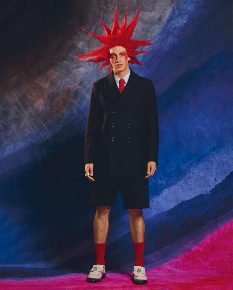 Young man in avant-garde fashion with red hair and dark overcoat, red socks and loafers posing against an abstract background.