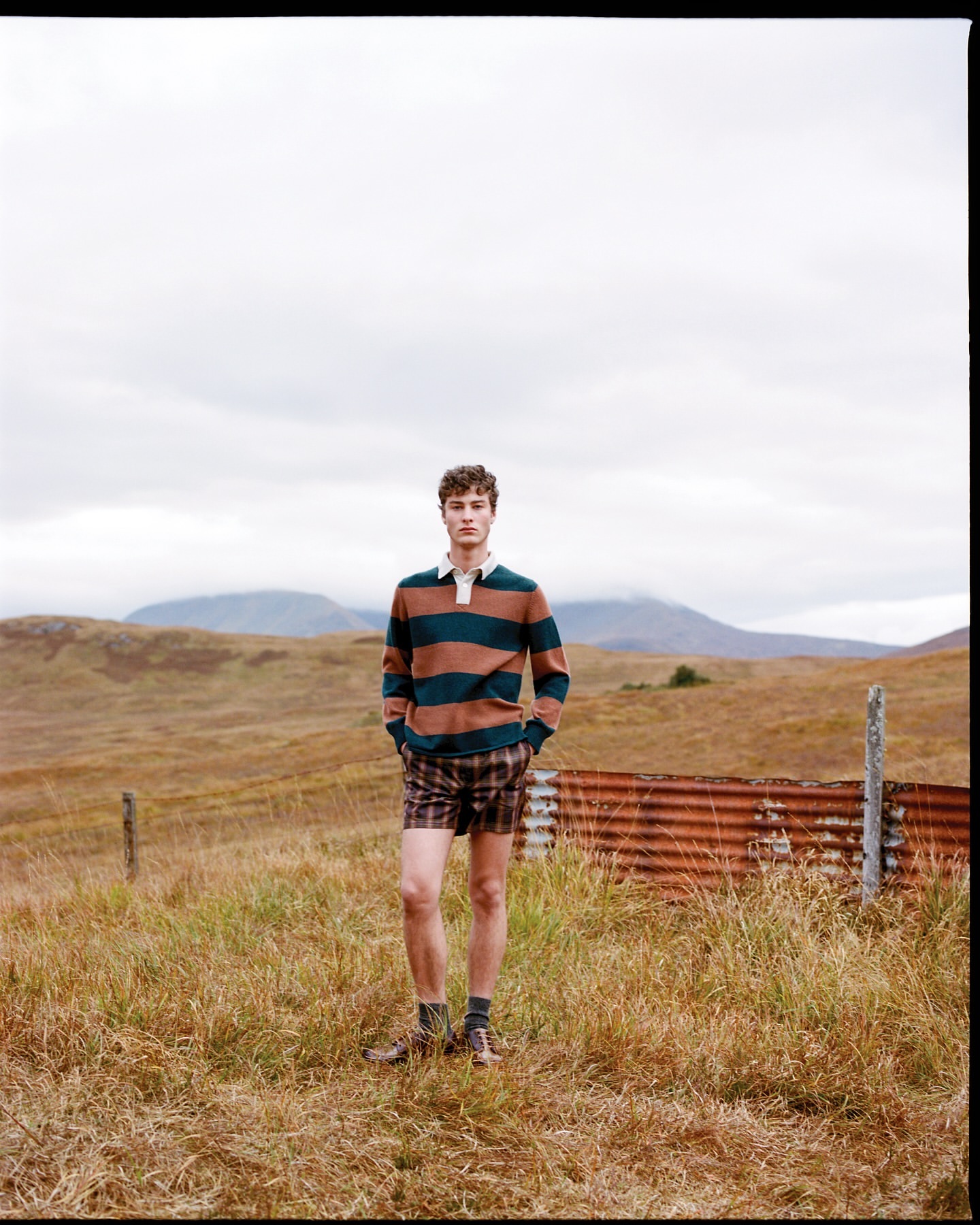 Adult male standing in a grassy field wearing a striped rugby shirt and plaid shorts with a rusty metal structure behind him.