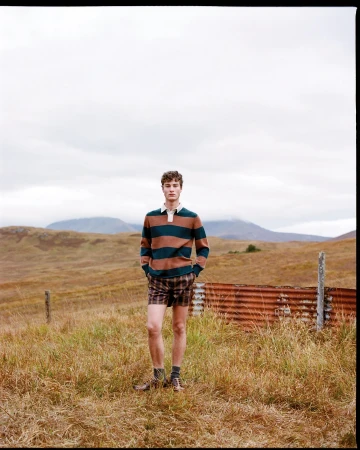 Adult male standing in a grassy field wearing a striped rugby shirt and plaid shorts with a rusty metal structure behind him.