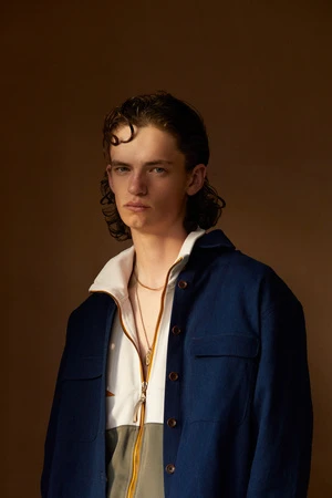 Portrait of a young man with dark wavy hair wearing a blue overshirt and zip-up jacket against a warm brown studio backdrop