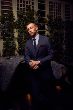 Formally dressed man in pinstripe suit poses indoors with greenery backdrop; fashion photography showcasing tailored menswear.