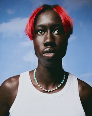 Portrait of a black man with vibrant red hair wearing a white tank top and beaded necklace against a blue sky background.