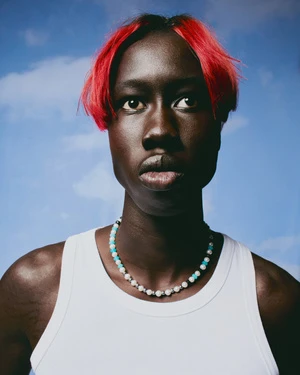 Portrait of a black man with vibrant red hair wearing a white tank top and beaded necklace against a blue sky background.