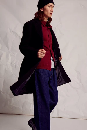 A long-haired person in a black beanie, dark coat, red shirt, and jeans poses against a textured white backdrop in a fashion editorial shot.