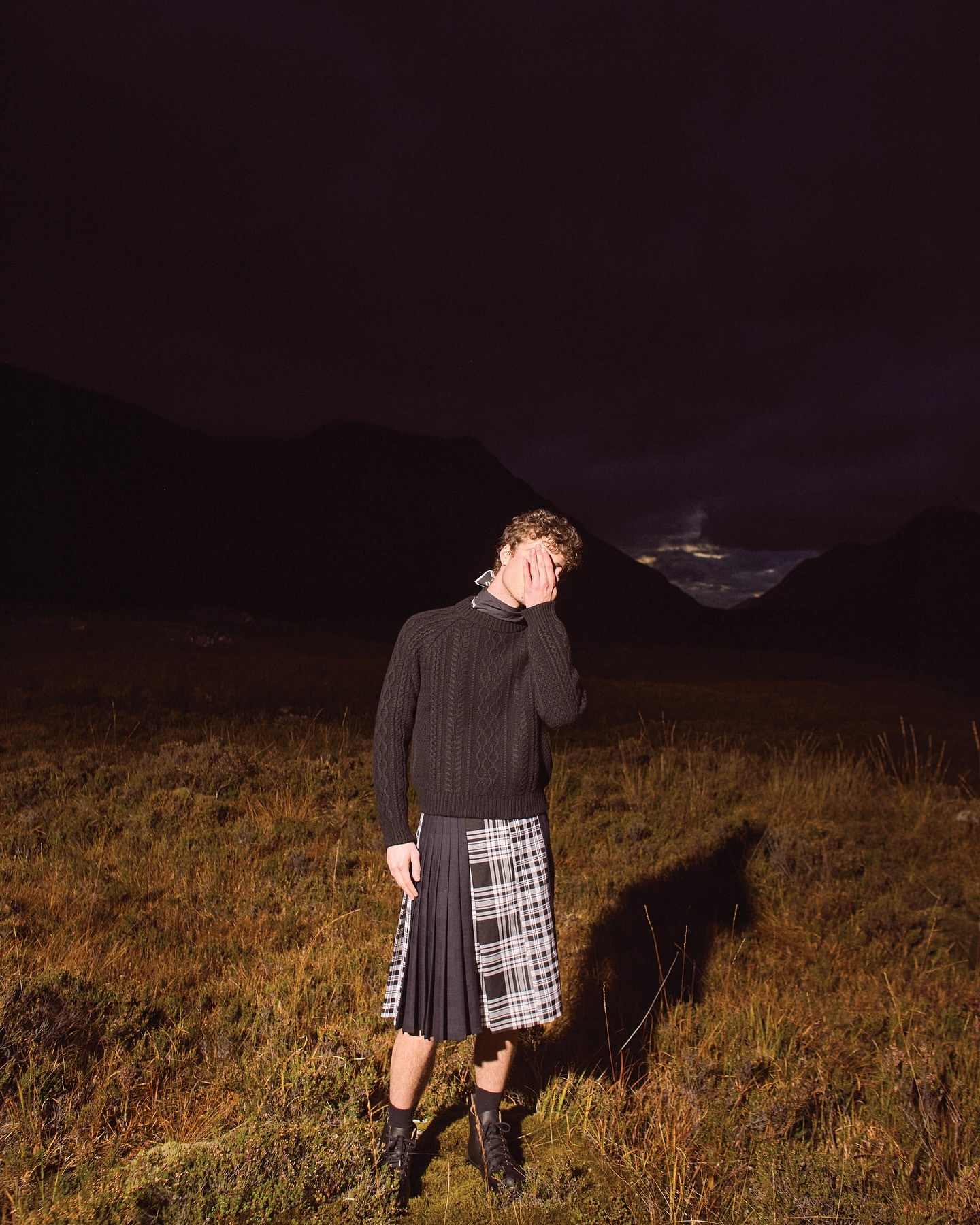 Man in kilt and sweater standing in a grassy field at night with mountains in the background. He has his hand to his face.
