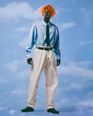 Full-length portrait of a Black man with bright orange hair wearing a light blue button-down shirt, dark tie, white trousers, and black shoes.