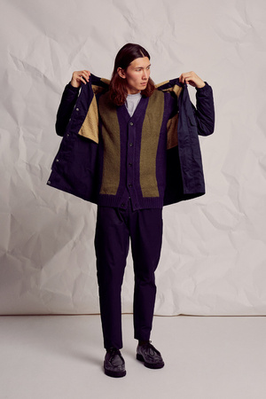 Full-shot of a male model posing in a blue jacket, cardigan, and navy pants against a textured off-white background in a studio setting.