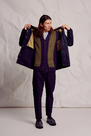 Full-shot of a male model posing in a blue jacket, cardigan, and navy pants against a textured off-white background in a studio setting.