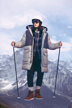 A male model in winter apparel poses with ski poles in front of a mountain range, wearing a helmet, goggles, and a large puffer coat.