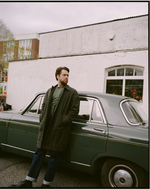 A man leans against a classic green sedan in an urban setting, wearing a dark green overcoat and jeans, under an overcast sky.