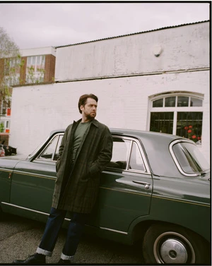 A man leans against a classic green sedan in an urban setting, wearing a dark green overcoat and jeans, under an overcast sky.