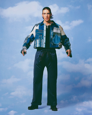 Full-length shot of a male model wearing a custom patchwork denim jacket and navy trousers, posing against a cloudy blue sky background.