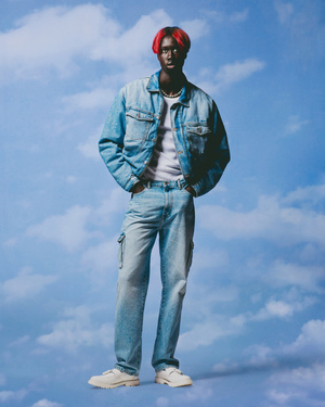 Black man with dyed red hair wearing a denim jacket and jeans against a sky background