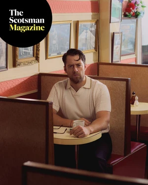 Adult male sits at table in diner with coffee and eyeglasses wearing collared shirt in medium shot, natural lighting, pensive expression.