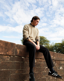 Young man sitting on a brick wall wearing a textured beige sweater, dark trousers, and black sneakers against a cloudy sky