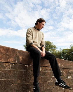 Young man sitting on a brick wall wearing a textured beige sweater, dark trousers, and black sneakers against a cloudy sky