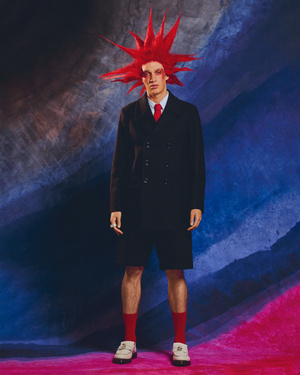 A man stands in a studio wearing a dark overcoat, shorts, and socks with a bold red hairstyle against a textured backdrop.