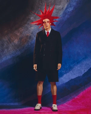 A man stands in a studio wearing a dark overcoat, shorts, and socks with a bold red hairstyle against a textured backdrop.