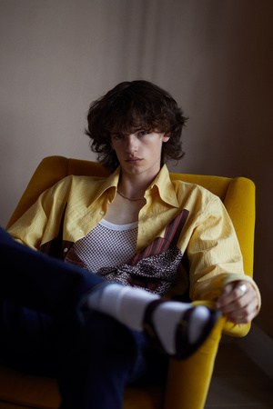 Stylish young adult sits in a yellow chair wearing a yellow shirt and white socks indoors, posing with a neutral expression.