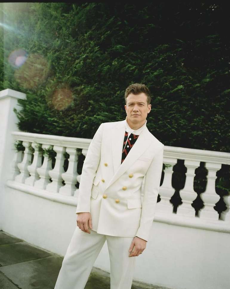Man in a cream-colored suit poses outdoors against a white wall and green hedge in an eye-level, medium shot with natural lighting.