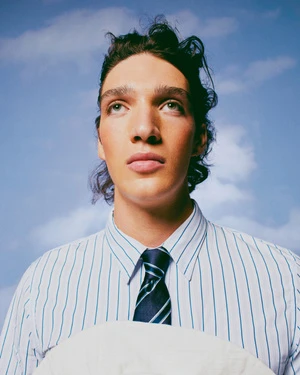 Close-up portrait of a man with dark curly hair wearing a blue pinstripe shirt and tie against a cloudy blue sky background.