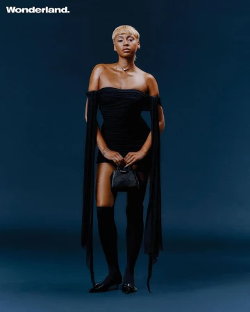 Model with blonde short hair wearing a black off-the-shoulder mini dress holding a small black handbag against a dark blue background.