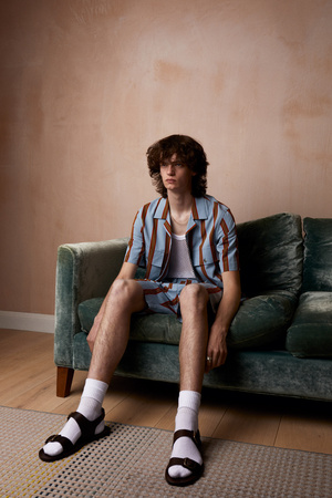 A young adult male with curly hair sitting on a green velvet couch wearing a striped shirt and shorts with black sandals indoors.
