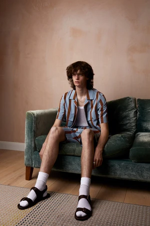 A young adult male with curly hair sitting on a green velvet couch wearing a striped shirt and shorts with black sandals indoors.