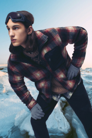 An adult male poses in the outdoors. He is wearing a plaid jacket, dark pants, gloves, and ski goggles pushed back on his head.